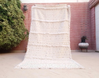 May include: A large, rectangular, off-white area rug featuring a thick, shaggy texture with horizontal rows of fringe detailing. The surface is embellished with small, reflective, circular metallic accents scattered across the woven fabric.