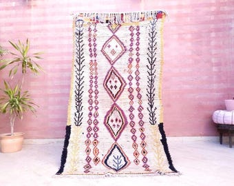 Puede incluir: Una alfombra rectangular con base de color crema que presenta una columna central vertical de cuatro grandes formas de diamante en tonos magenta, negro y naranja. La alfombra incluye patrones geométricos intrincados, líneas y flecos.