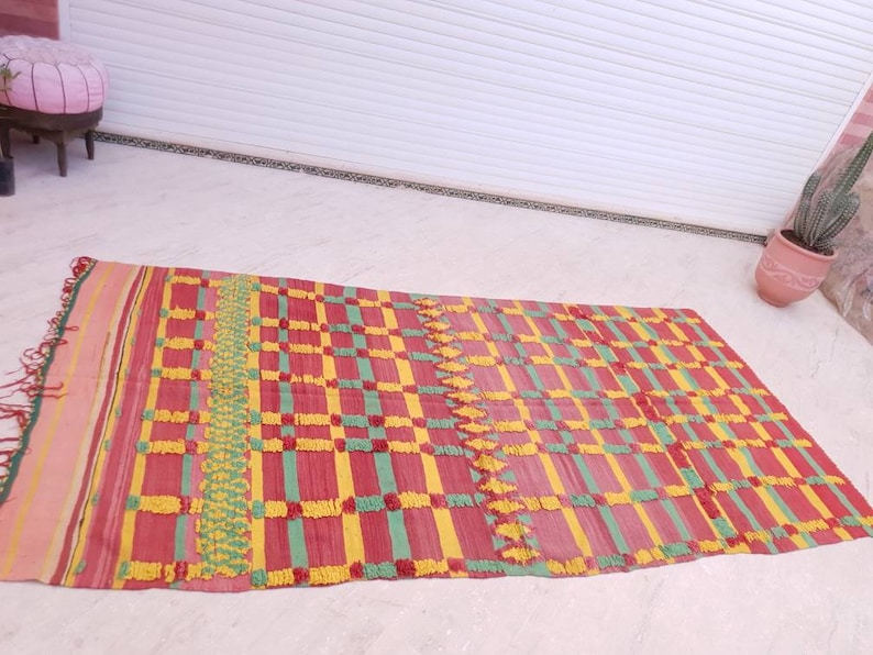May include: A rectangular woven rug with a checkered pattern in shades of red, yellow, and green. The rug has a fringe border and a textured surface.