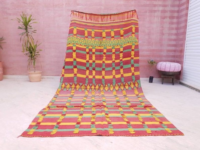 May include: A colorful Moroccan rug with a geometric pattern in shades of red, green, and yellow. The rug has a woven texture and is made of wool.