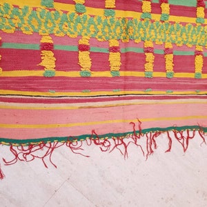 May include: A colorful woven rug with a geometric pattern in shades of pink, yellow, green, and black. The rug has a fringe border.