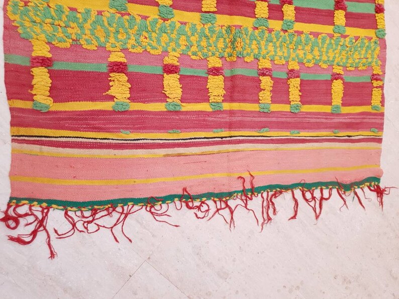 May include: A colorful woven rug with a geometric pattern in shades of pink, yellow, green, and black. The rug has a fringe border.