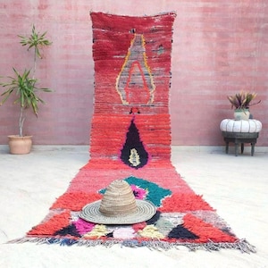 May include: A long, rectangular runner rug featuring a vibrant red base with a textured, shaggy pile. The rug displays abstract geometric patterns in black, teal, yellow, and pink, and is topped with a woven straw hat with a wide brim and pointed crown.