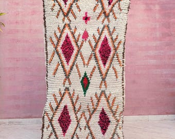 May include: A rectangular, off-white wool rug featuring a high-pile texture and a repeating geometric diamond pattern. The design incorporates vibrant magenta, orange, and dark green accents within the diamonds, finished with a dark brown border.