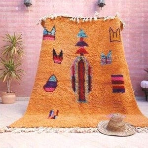 May include: A large, rectangular, orange shag rug featuring a central vertical column of stacked geometric shapes in red, blue, and purple. The rug is decorated with scattered, colorful abstract motifs and has a fringed edge at the top and bottom.