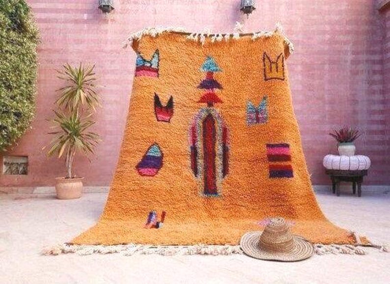 May include: A large, rectangular, orange shag rug featuring a central vertical column of stacked geometric shapes in red, blue, and purple. The rug is decorated with scattered, colorful abstract motifs and has a fringed edge at the top and bottom.