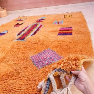 May include: A rectangular orange shag rug featuring scattered geometric shapes and abstract patterns in blue, pink, purple, and red. The rug has a thick, textured pile and includes braided fringe tassels along the edge with white yarn accents.