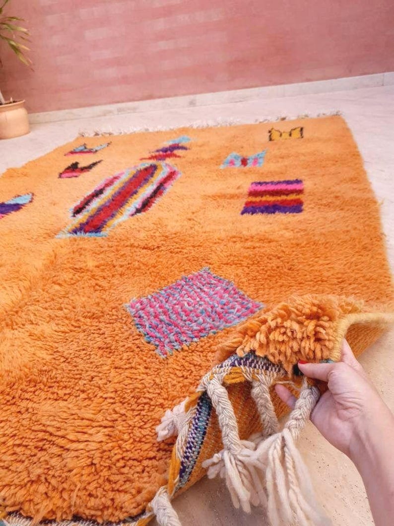 May include: A rectangular orange shag rug featuring scattered geometric shapes and abstract patterns in blue, pink, purple, and red. The rug has a thick, textured pile and includes braided fringe tassels along the edge with white yarn accents.