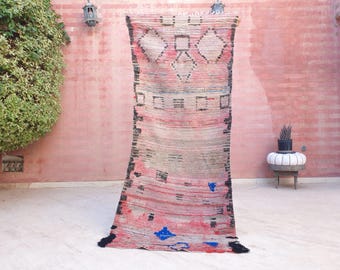 Puede incluir: Una alfombra tejida rectangular con una base de color rosa y beige descolorido, con bandas horizontales de patrones geométricos en negro, azul y blanco, incluyendo diamantes y cuadrados. Los bordes están terminados con flecos negros y bordes verticales oscuros.