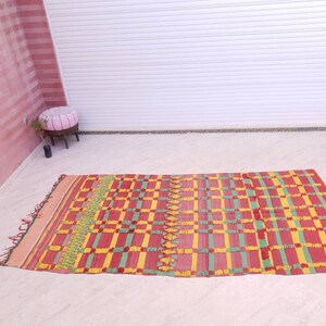 May include: A red, yellow, and green patterned rug with fringe. The rug has a checkered design with a geometric pattern.