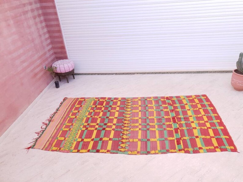 May include: A red, yellow, and green patterned rug with fringe. The rug has a checkered design with a geometric pattern.
