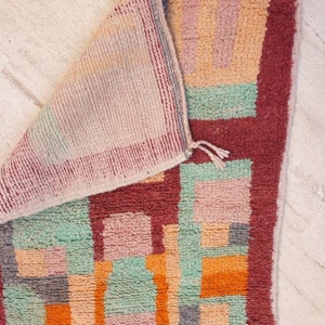 May include: A colorful geometric rug with a red border. The rug is made of wool and has a hand-woven texture. The colors are pink, orange, green, and gray.