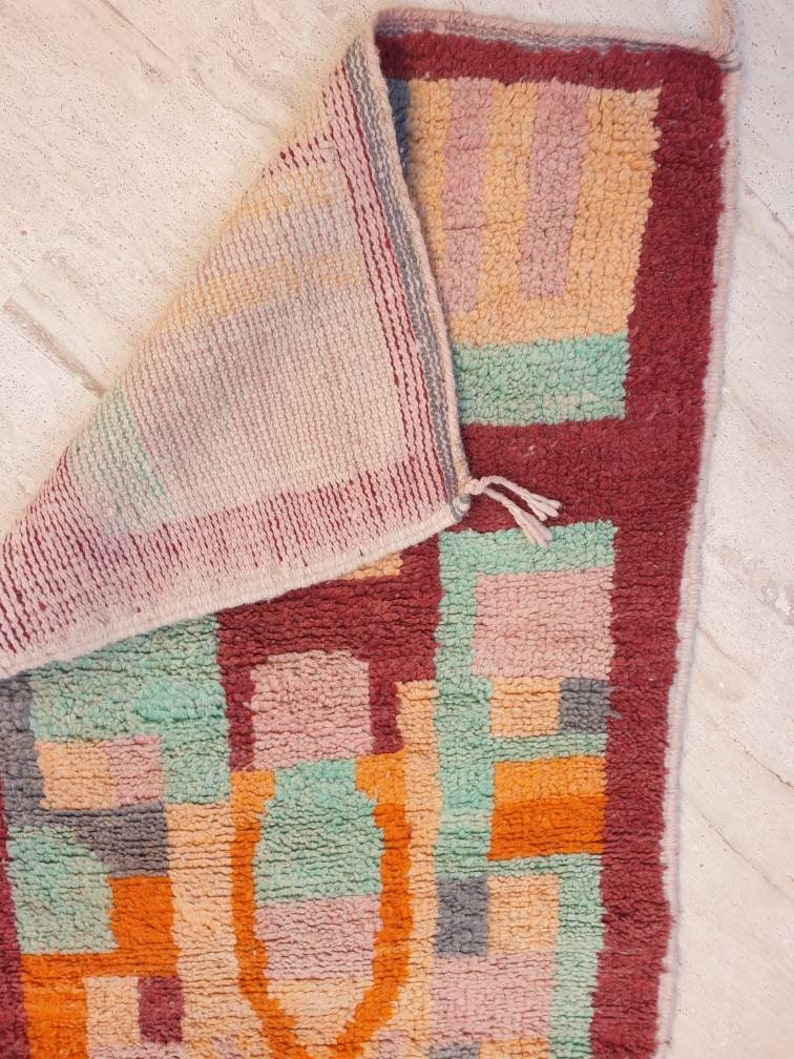 May include: A colorful geometric rug with a red border. The rug is made of wool and has a hand-woven texture. The colors are pink, orange, green, and gray.