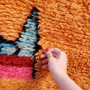 May include: A thick, high-pile shag rug featuring a vibrant orange base color. The rug displays a geometric pattern with sections of bright blue, deep red, and hot pink yarn, all outlined with bold black fibers throughout the textured surface.