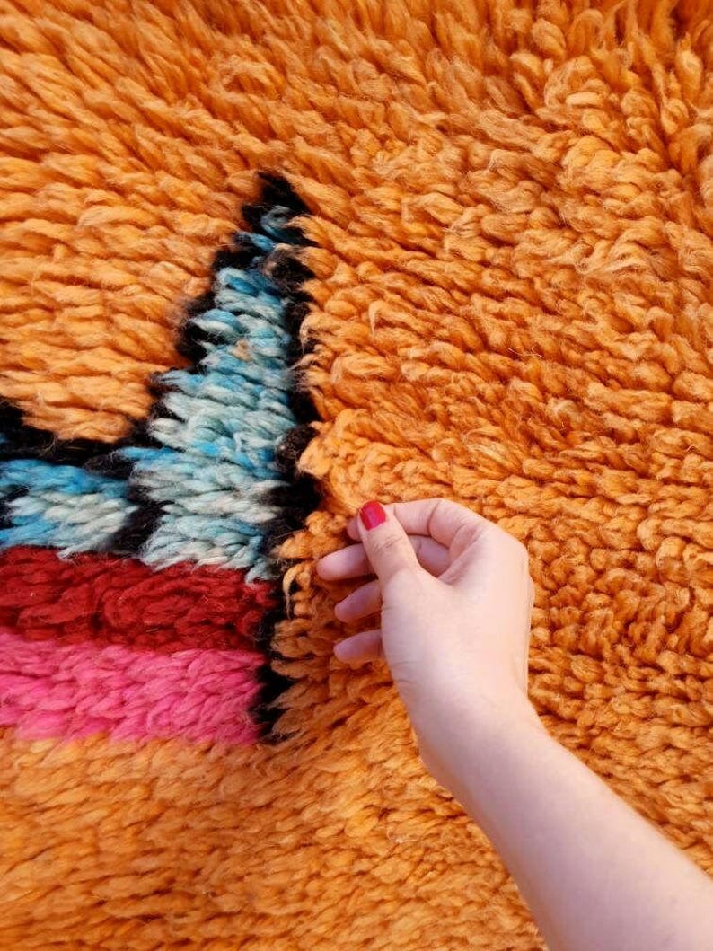May include: A thick, high-pile shag rug featuring a vibrant orange base color. The rug displays a geometric pattern with sections of bright blue, deep red, and hot pink yarn, all outlined with bold black fibers throughout the textured surface.