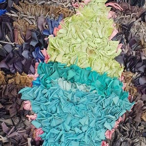 Puede incluir: Una alfombra colorida hecha de recortes de tela reciclados. La alfombra presenta una forma de diamante en tonos de verde, azul y rosa.