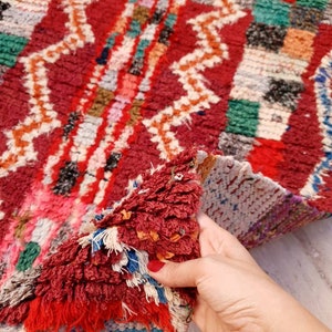 May include: A close-up of a colorful Moroccan rug with a hand holding the edge of the rug. The rug has a geometric pattern with shades of red, green, white, and blue.