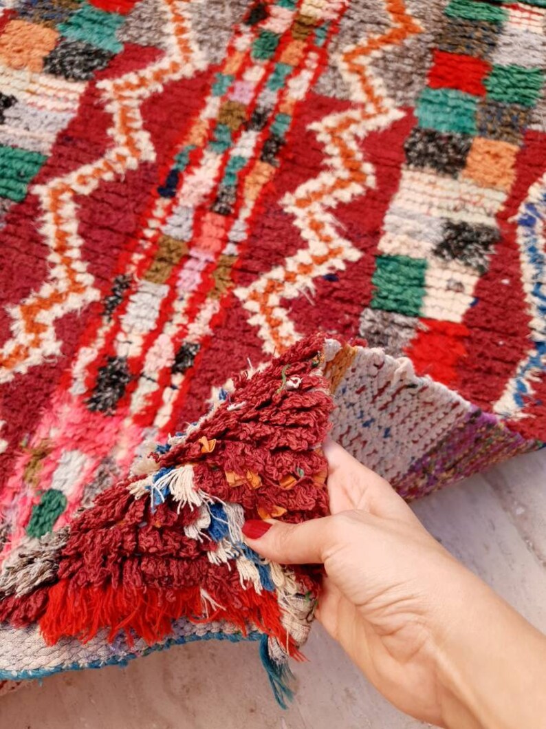 May include: A close-up of a colorful Moroccan rug with a hand holding the edge of the rug. The rug has a geometric pattern with shades of red, green, white, and blue.