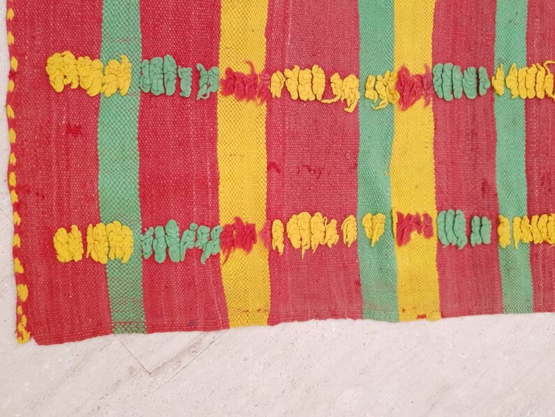May include: A woven rug with red, yellow, and green stripes. The rug has a textured surface with raised loops in yellow and green.