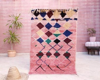 Puede incluir: Una alfombra marroquí rosa con un patrón de diamantes en tonos de azul, verde y marrón. La alfombra tiene un borde de flecos y está tejida con una variedad de texturas.