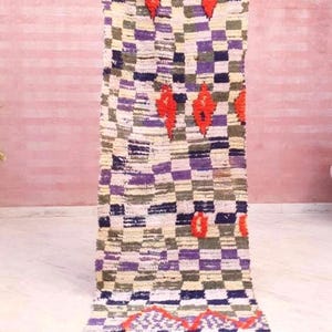 May include: A long rectangular runner rug featuring a checkered pattern in shades of purple, dark blue, olive green, and cream. The rug is accented with bright orange diamond-shaped motifs and a zigzag line near the bottom, finished with fringe.