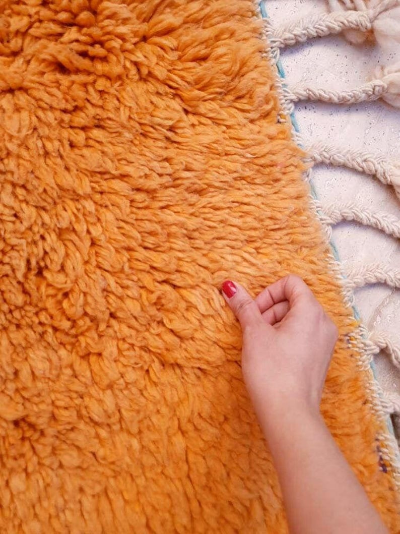 May include: A thick, high-pile rug in a vibrant orange hue featuring a dense, shaggy texture. The edge of the rug is finished with a series of cream-colored, braided fringe tassels that extend outward from the main body of the textile piece.