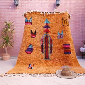May include: A large, rectangular, orange wool rug featuring a central vertical totem-like pattern in red, blue, and purple, surrounded by scattered geometric shapes in various colors. The rug has a thick, shaggy texture and white fringe at both ends.
