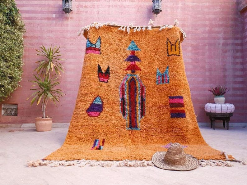 May include: A large, rectangular, orange wool rug featuring a central vertical totem-like pattern in red, blue, and purple, surrounded by scattered geometric shapes in various colors. The rug has a thick, shaggy texture and white fringe at both ends.