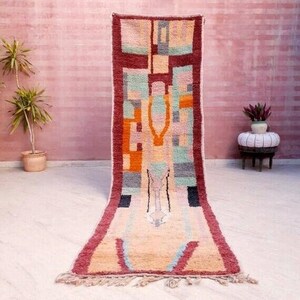 May include: A long, rectangular rug with a geometric pattern in shades of red, orange, green, blue, and beige. The rug has a fringe on the ends.