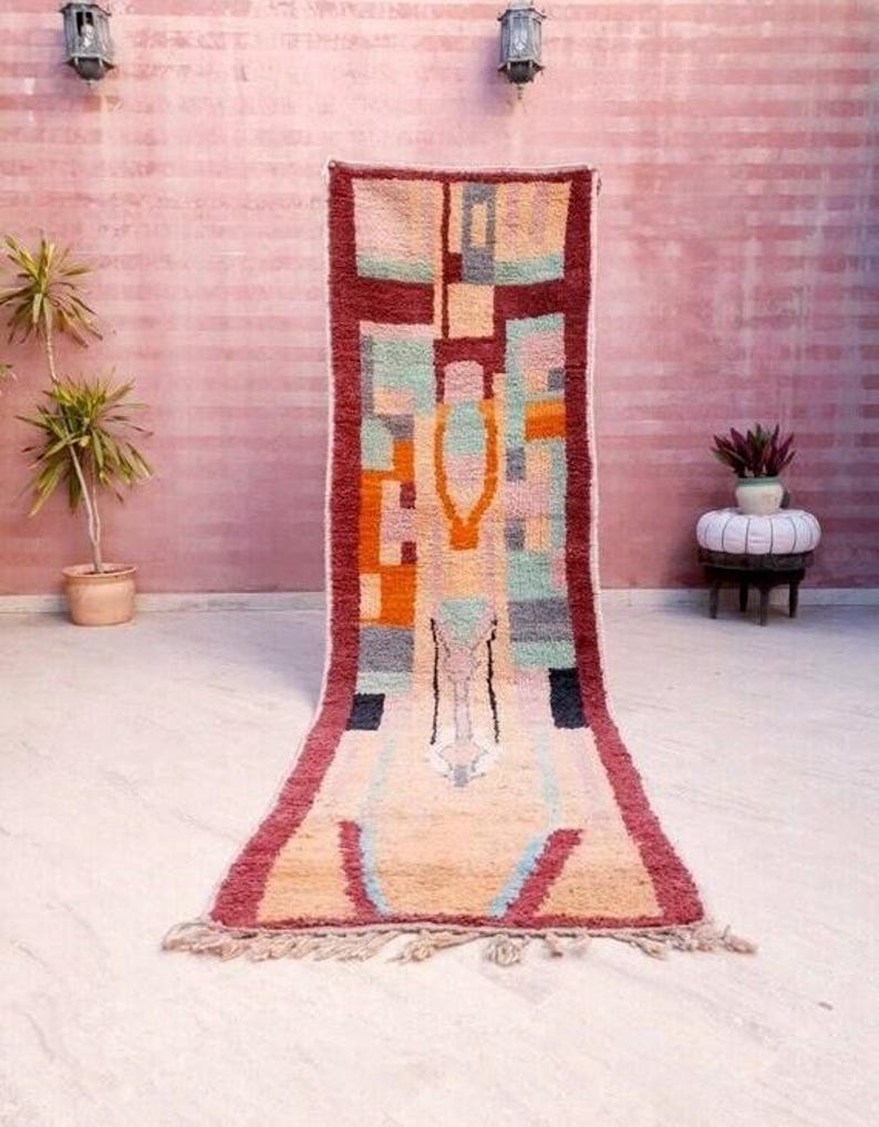 May include: A long, rectangular rug with a geometric pattern in shades of red, orange, green, blue, and beige. The rug has a fringe on the ends.