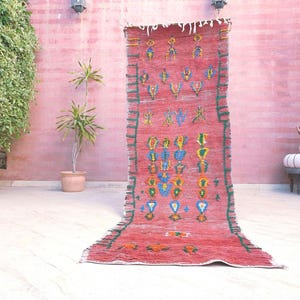 May include: A long, rectangular runner rug featuring a vibrant red base color with a series of small, colorful geometric patterns in blue, yellow, and green. The rug has fringed edges at the top and dark green stitched borders along the sides.