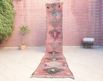 Puede incluir: Una alfombra de pasillo larga y estrecha con una base de color rosa vibrante y una serie de patrones geométricos en forma de diamante en gris oscuro y negro a lo largo del centro. La alfombra tiene un aspecto tejido y texturizado con flecos.