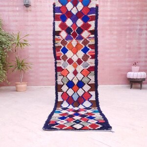 May include: A long, rectangular runner rug featuring a repeating diamond pattern in vibrant colors including red, blue, orange, purple, and brown against a cream-colored background, finished with dark blue borders along the long edges.
