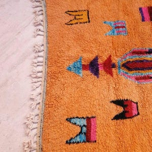 May include: A rectangular orange area rug featuring a high-pile texture and a series of colorful geometric motifs in shades of blue, purple, pink, and black. The rug is finished with a row of off-white knotted fringe along the left edge.