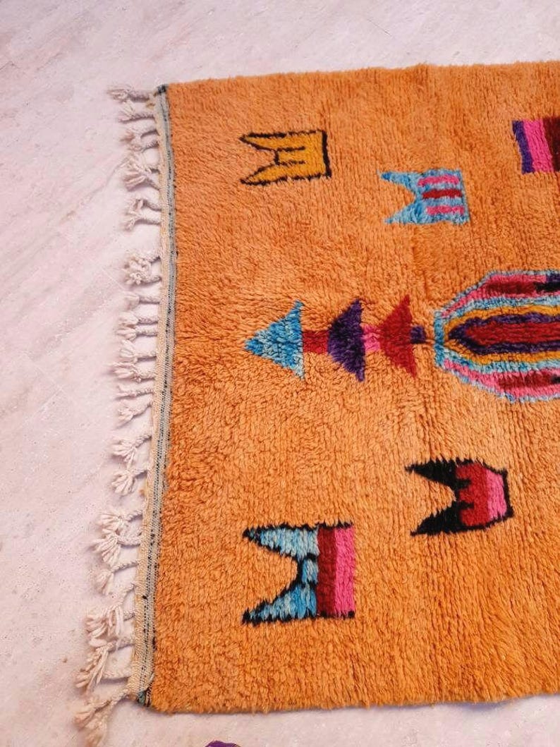 May include: A rectangular orange area rug featuring a high-pile texture and a series of colorful geometric motifs in shades of blue, purple, pink, and black. The rug is finished with a row of off-white knotted fringe along the left edge.