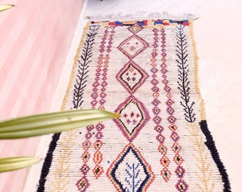 May include: A white Moroccan rug with a geometric pattern in shades of red, blue, yellow, and black. The rug has a fringe border and is laid out on a tiled floor.