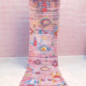 May include: A long, rectangular runner rug featuring a vibrant pink base with horizontal stripes and scattered, multi-colored geometric patterns in shades of blue, yellow, and red. The rug has a textured, woven surface and frayed fringe edges.