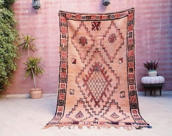 May include: A large, rectangular rug with a peach-colored background and intricate geometric patterns in black and red. The rug features a diamond-shaped central design and a border with repeating shapes. The rug is displayed outdoors.