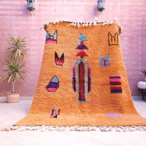 May include: A rectangular orange area rug featuring a high-pile texture and a central vertical column design. The rug is decorated with scattered geometric shapes in blue, purple, pink, and black, and includes white fringe along the top and bottom edges.
