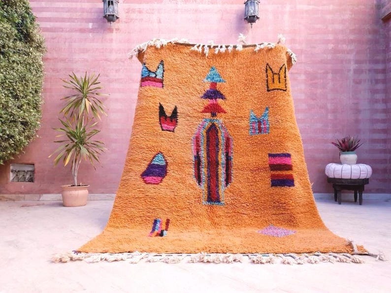 May include: A rectangular orange area rug featuring a high-pile texture and a central vertical column design. The rug is decorated with scattered geometric shapes in blue, purple, pink, and black, and includes white fringe along the top and bottom edges.
