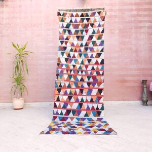May include: A long, rectangular runner rug featuring a repeating geometric pattern of small triangles in vibrant colors including orange, purple, green, black, and white, arranged in horizontal rows across the entire length of the textile.