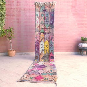 Puede incluir: Una alfombra de pasillo larga y estrecha con un vibrante patrón geométrico multicolor. El diseño incluye motivos de árboles estilizados, diamantes y formas de cruz en tonos rosa, amarillo, morado, turquesa y gris, con un borde de flecos.