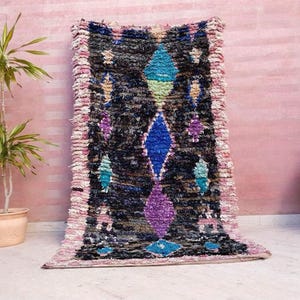 Puede incluir: Una alfombra marroqu&iacute; colorida con un patr&oacute;n geom&eacute;trico en tonos de azul, rosa, morado y verde. La alfombra tiene un borde con flecos y est&aacute; hecha de lana.