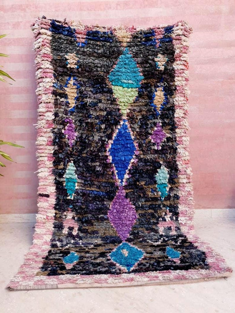 Puede incluir: Una alfombra marroqu&iacute; colorida con un patr&oacute;n geom&eacute;trico en tonos de azul, verde, morado y rosa. La alfombra tiene un aspecto desgastado y vintage y est&aacute; hecha de lana.
