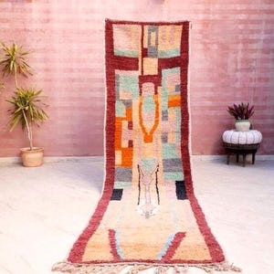 May include: A long, rectangular rug with a geometric pattern in shades of red, orange, yellow, green, blue, and gray. The rug has a fringe border and is hanging on a pink wall.