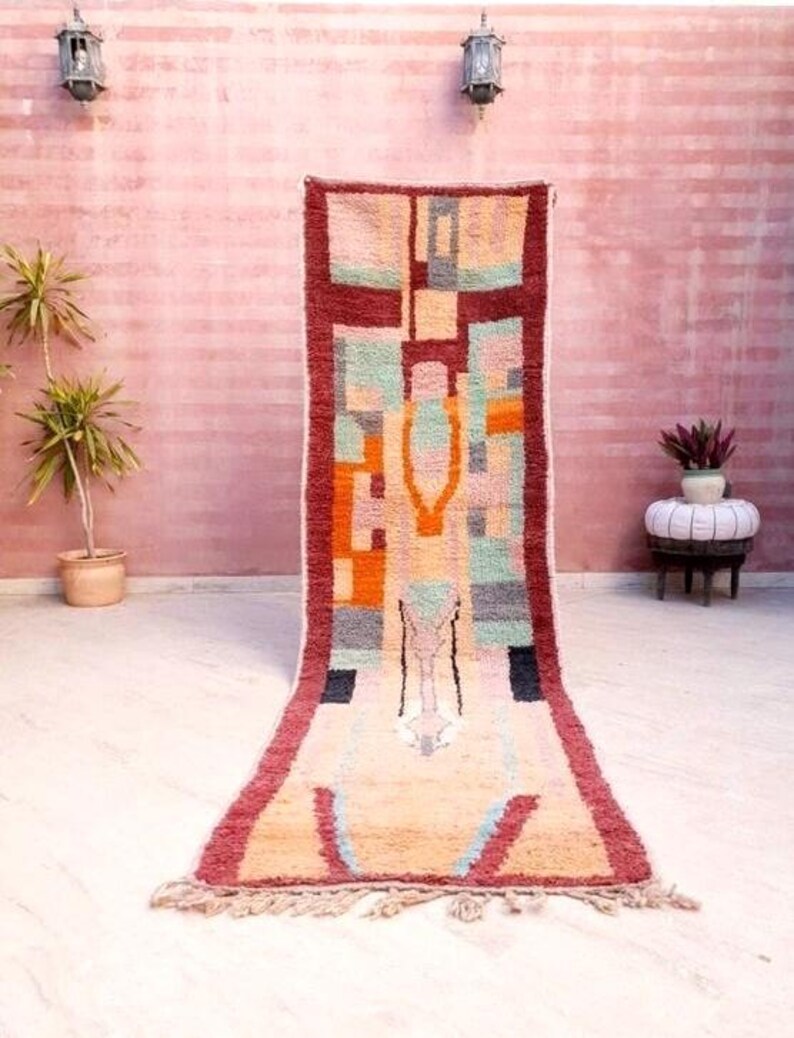 May include: A long, rectangular rug with a geometric pattern in shades of red, orange, yellow, green, blue, and gray. The rug has a fringe border and is hanging on a pink wall.