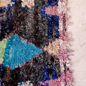 Puede incluir: Una alfombra colorida tejida a mano con un dise&ntilde;o de patchwork. La alfombra est&aacute; hecha de varios trozos de tela de color azul, rosa, blanco y gris. La alfombra tiene un borde deshilachado y una superficie texturizada.