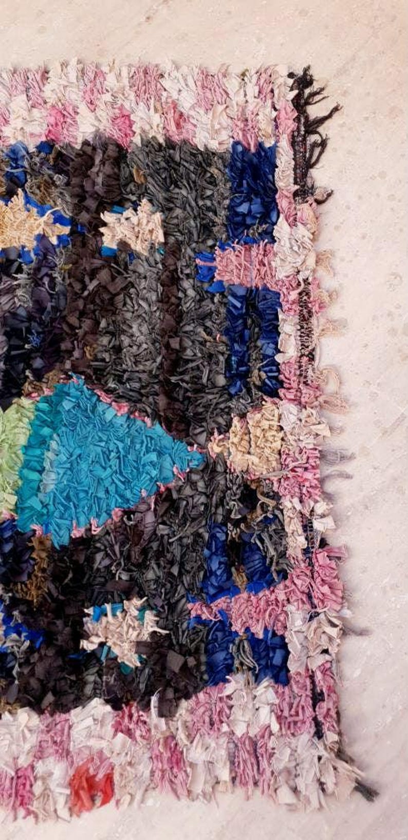 Puede incluir: Una alfombra colorida tejida a mano con un dise&ntilde;o de patchwork. La alfombra est&aacute; hecha de varios trozos de tela de color azul, rosa, blanco y gris. La alfombra tiene un borde deshilachado y una superficie texturizada.