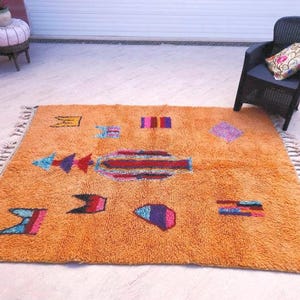 May include: A rectangular orange area rug featuring a high-pile texture and a series of scattered, multi-colored geometric shapes and symbols. The rug is finished with knotted fringe along two opposing edges, creating a textured border.