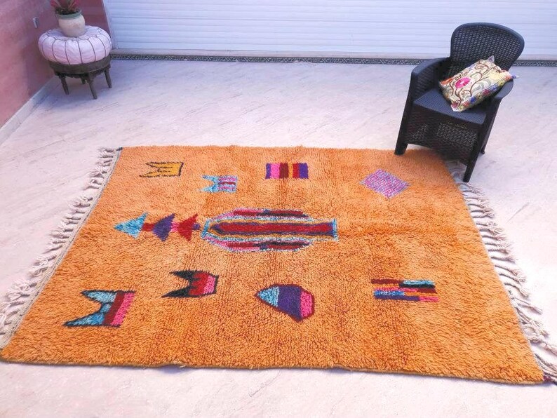 May include: A rectangular orange area rug featuring a high-pile texture and a series of scattered, multi-colored geometric shapes and symbols. The rug is finished with knotted fringe along two opposing edges, creating a textured border.